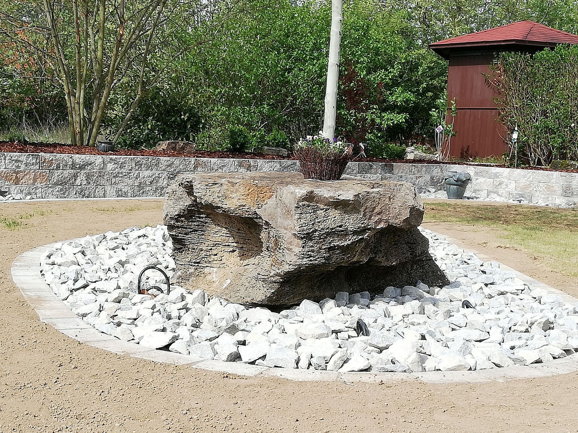 Steinbrunnen in einem Garten, umgeben von weißem Kies und einer geschwungenen Umrandung.