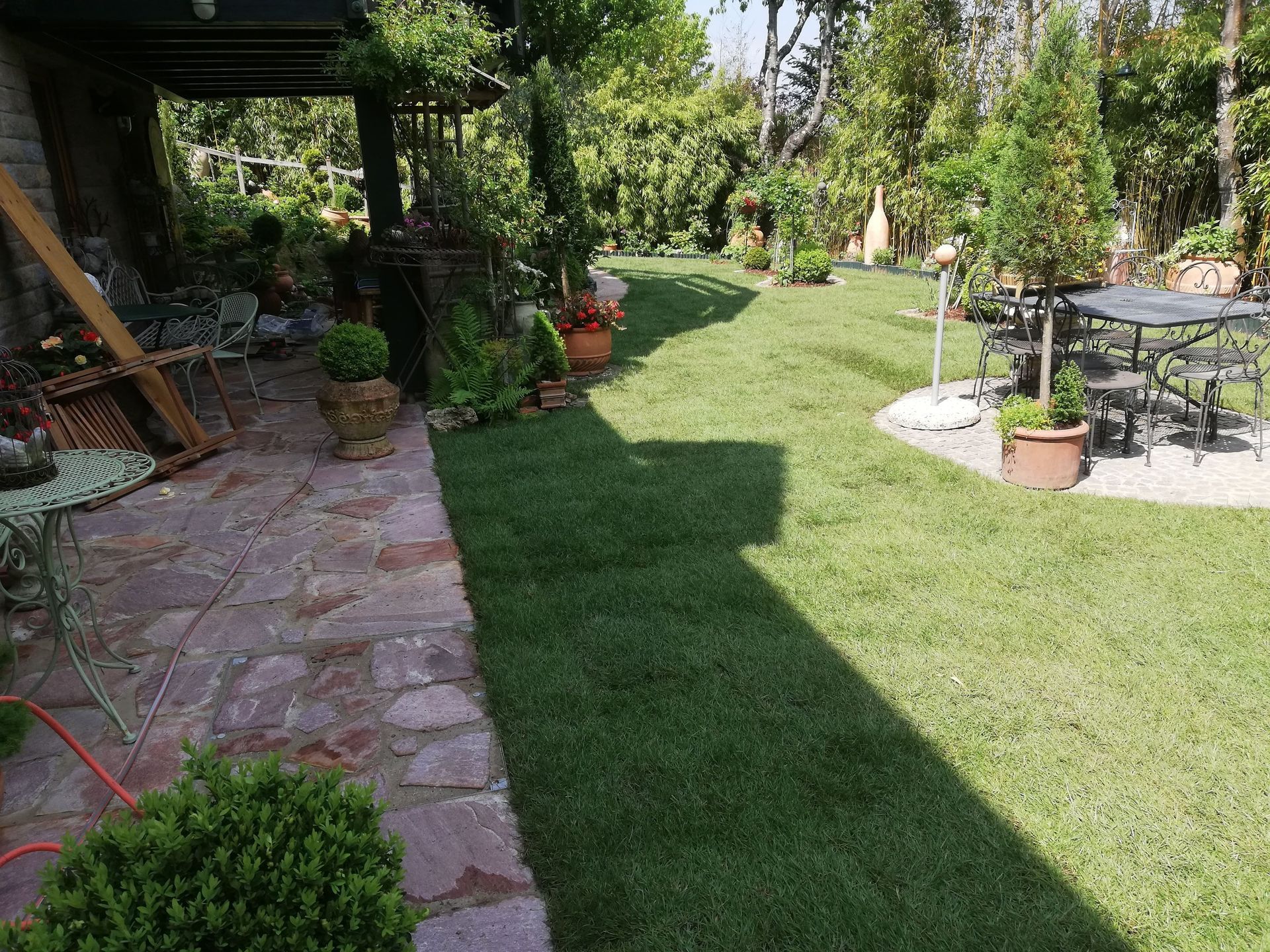 Grüner Rasen, gemauerte Terrasse mit Möbeln, umgeben von üppigen Bäumen in einem sonnigen Garten.