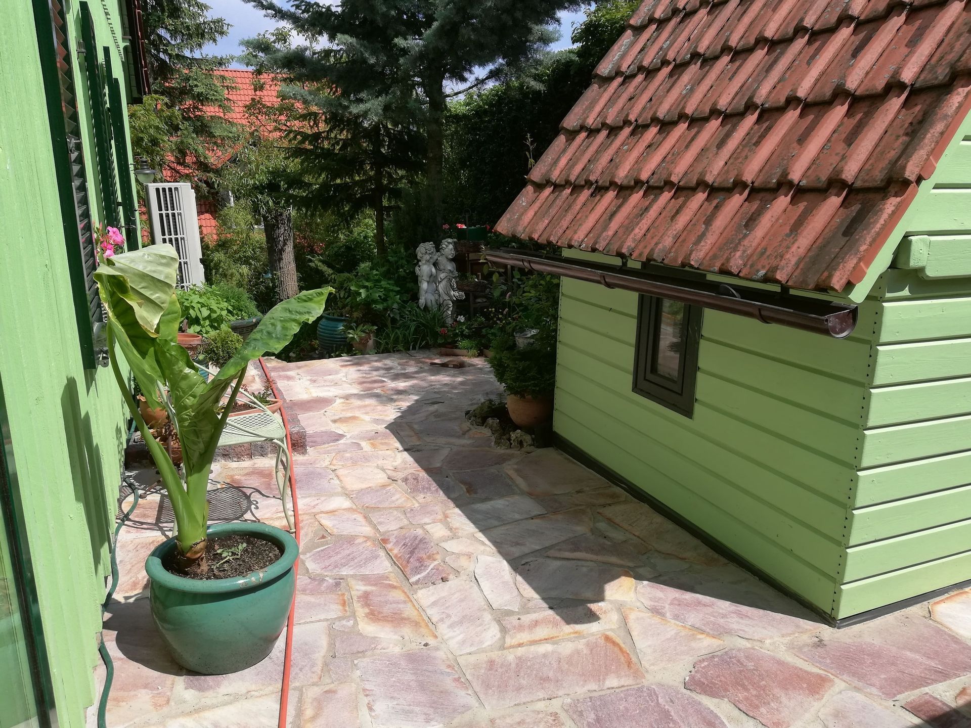 Haus mit grünen Seiten, rotem Dach und Steinweg, Topfpflanze im Vordergrund, Hinterhof-Einstellung.