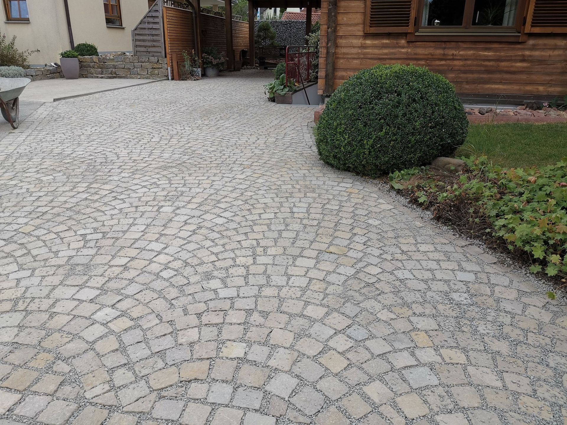 Die gepflasterte Auffahrt führt in Richtung eines Holzgebäudes, auf dessen rechter Seite sich ein runder grüner Busch befindet.