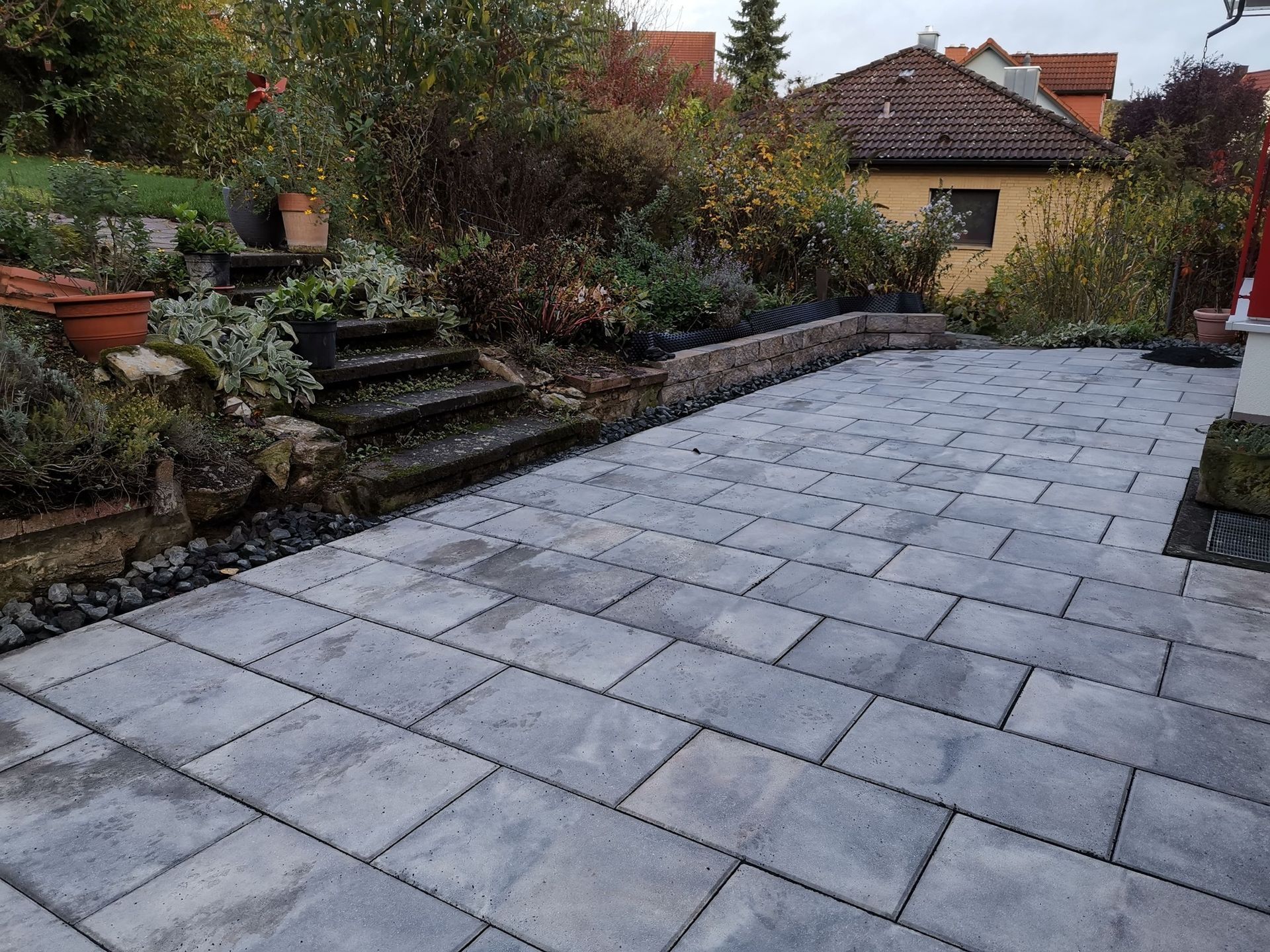 Mit grauen Blöcken gepflasterte Terrasse neben einem Garten mit Stufen und einem kleinen Gebäude.