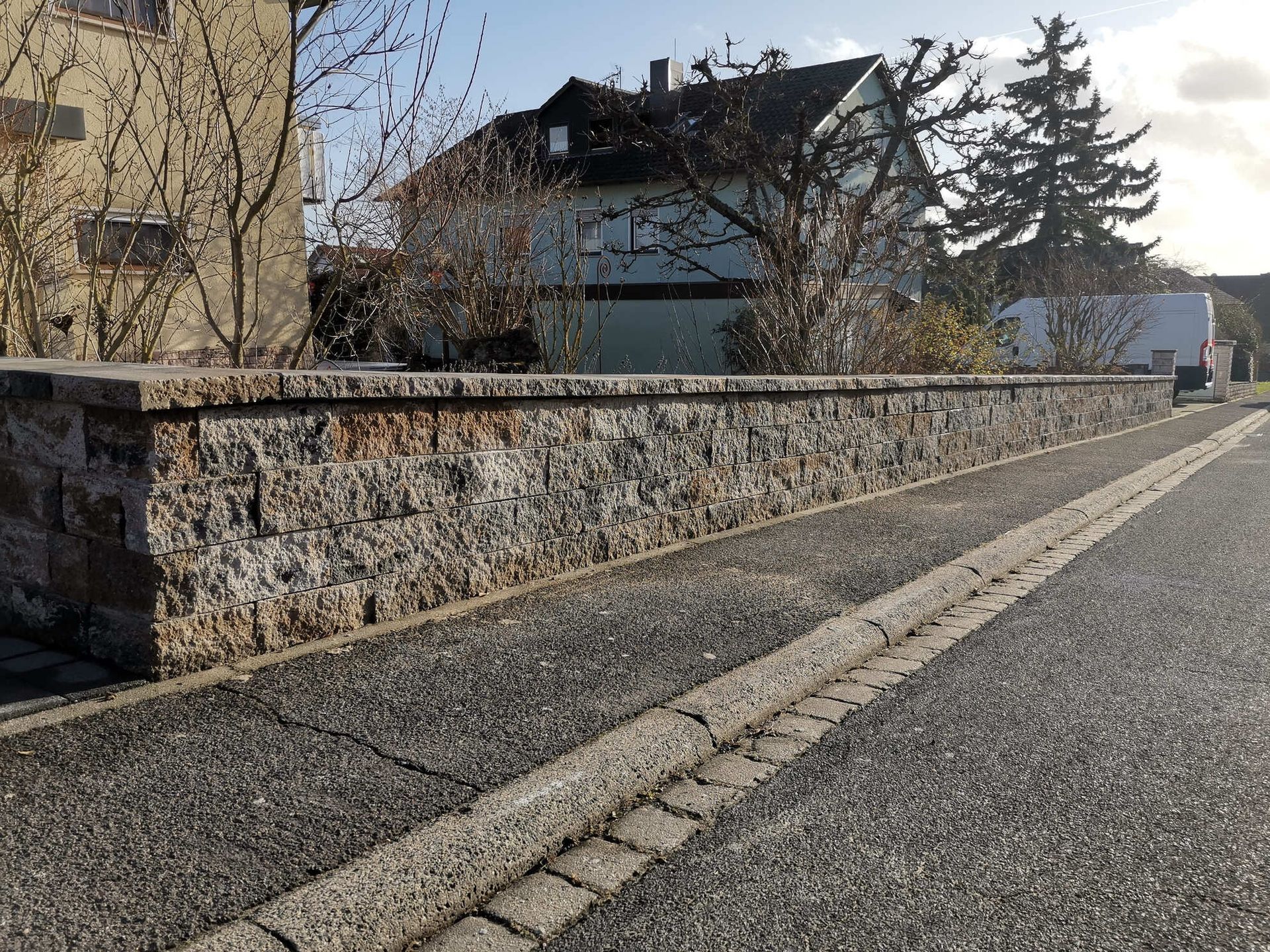 Steinmauer entlang einer Straße, mit einem Haus und Bäumen im Hintergrund.