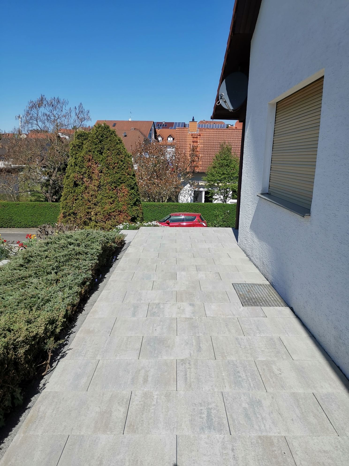 Weg mit Pflastersteinen, Hecke, Rasenmäher und Hausseite unter einem klaren, blauen Himmel.