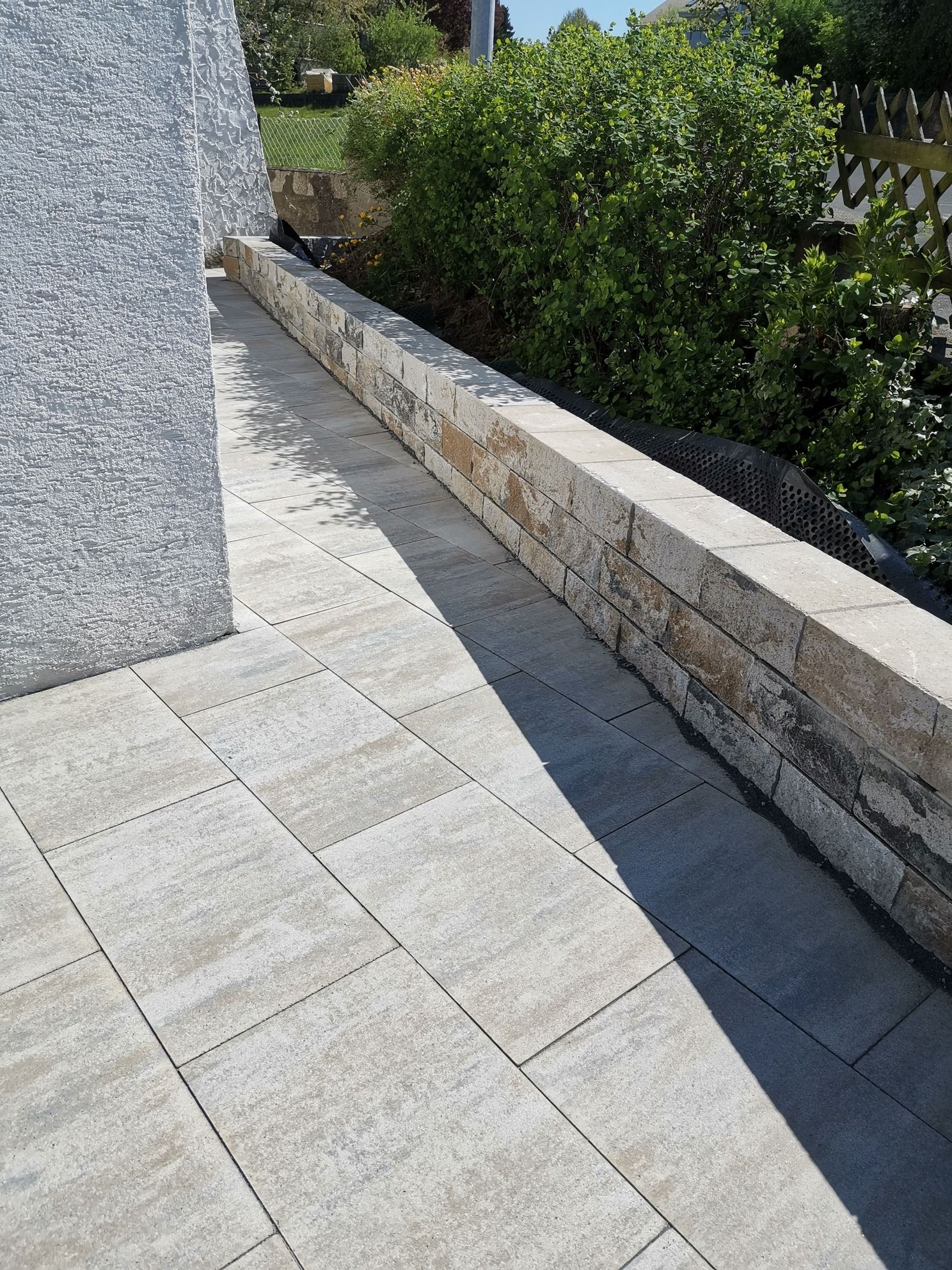 Steinterrasse mit niedriger Stützmauer, neben einer grauen Mauer und üppigem grünem Laub.