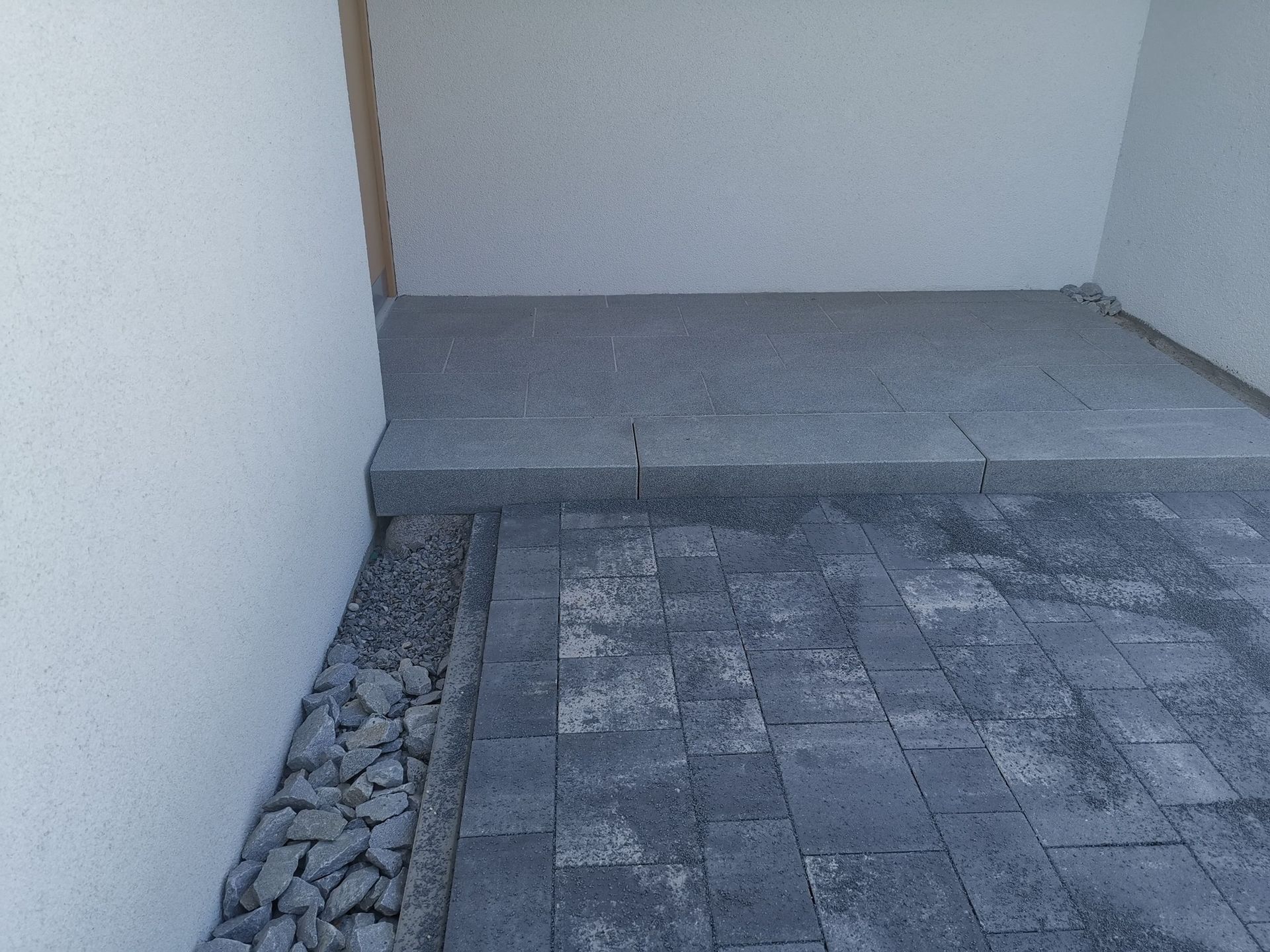 Grau gepflasterte Terrasse mit einer Stufe nach oben, begrenzt durch Felsen und weiße Mauern.