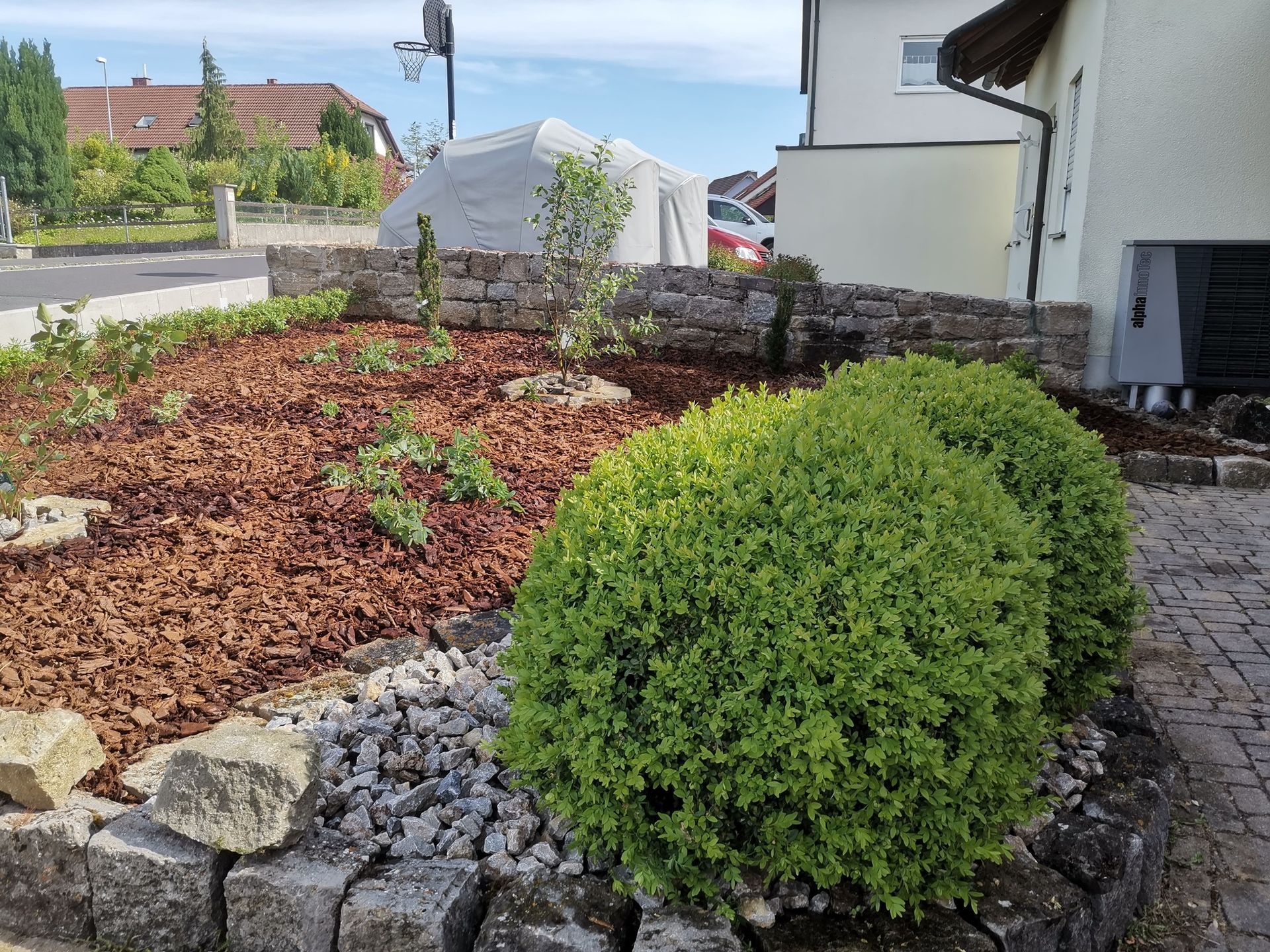 Landschaftsgärtnerischer Vorgarten mit grünen Büschen, braunem Mulch und einer Stützmauer aus Stein.