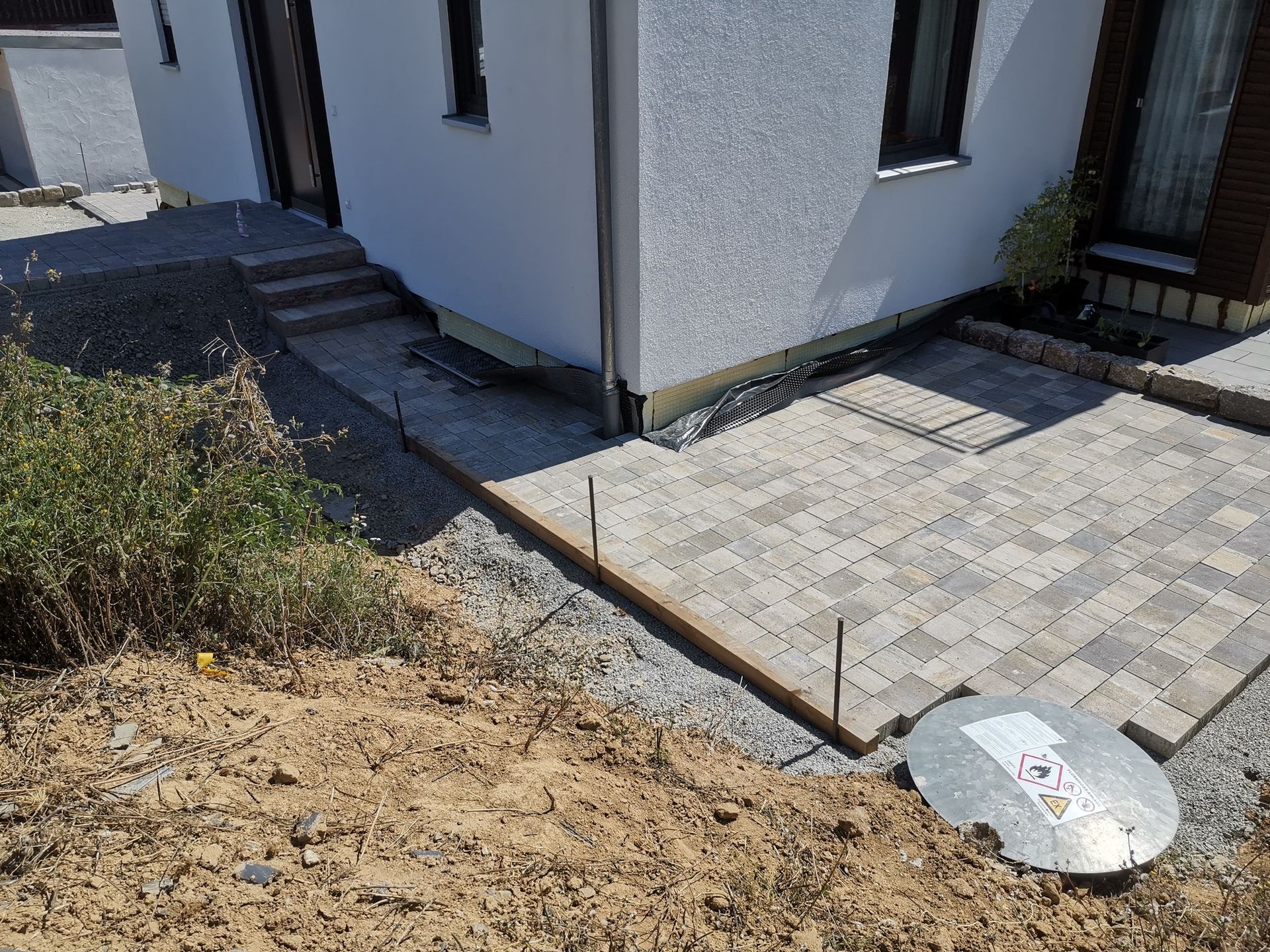 Eine teilweise angelegte Terrasse neben einem Haus. Pflastersteine, Kies und eine Stützmauer sind sichtbar.