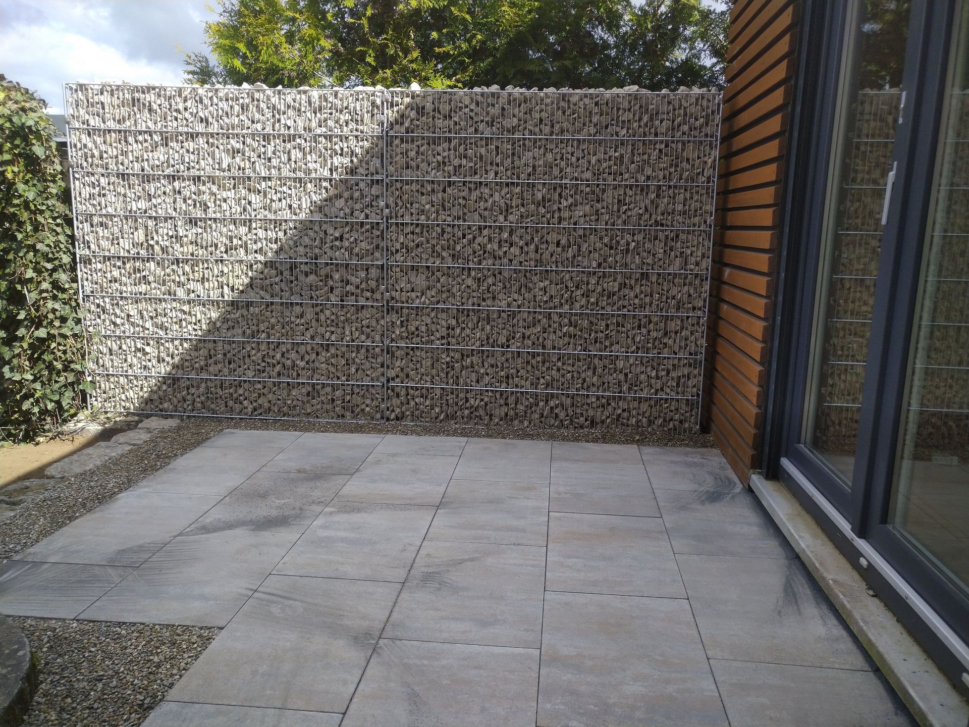 Terrasse mit Steinpflaster und einer mit Steinen gefüllten Gabionenwand.