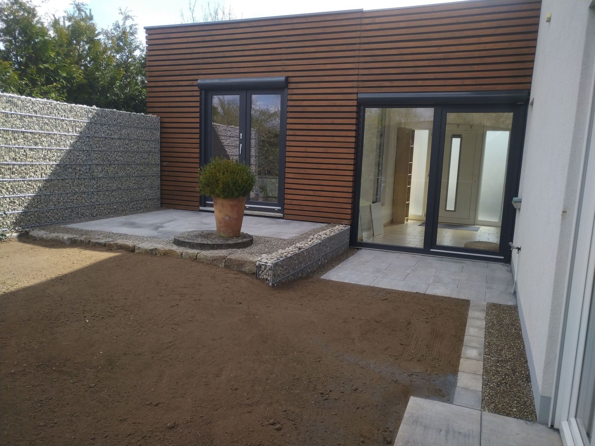 Außenansicht eines modernen Hauses mit brauner Holzverkleidung, Steinterrasse und einer Stützmauer aus Kies; in der Mitte steht eine Topfpflanze.