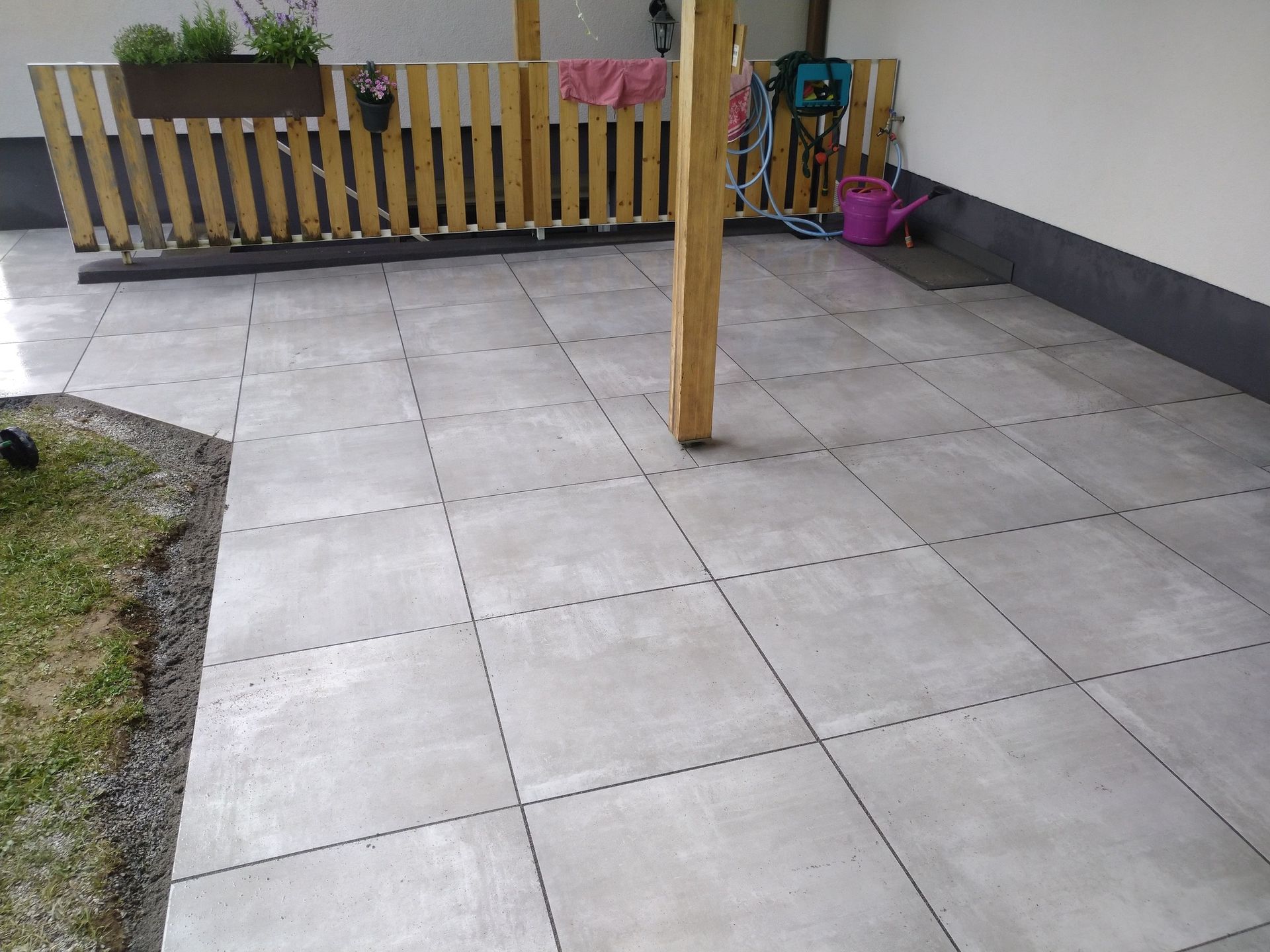 Grau geflieste Terrasse mit Holzzaun, Balken und Grasrand.