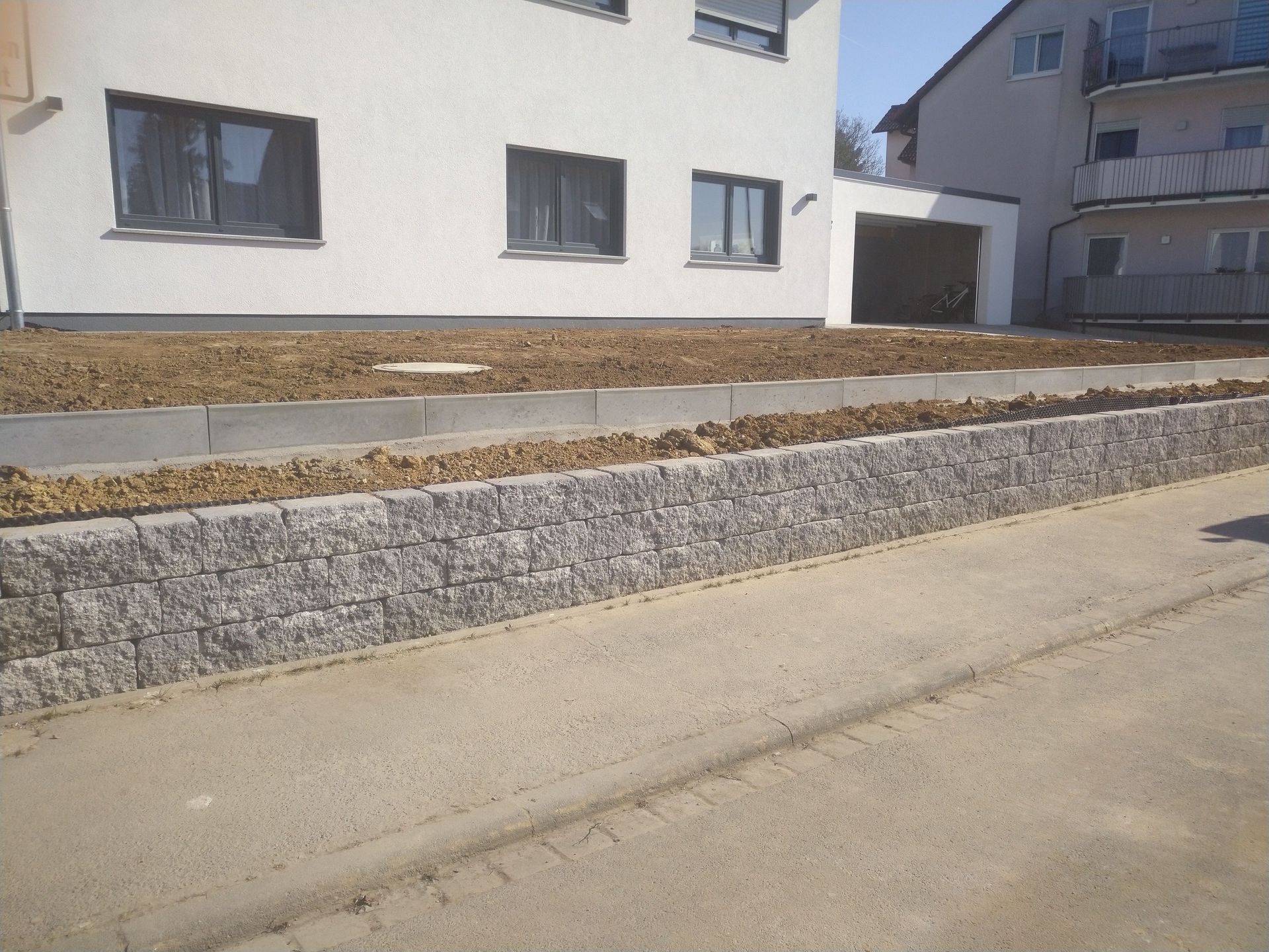 Eine niedrige Stützmauer aus grauem Stein vor einem weißen Gebäude, im Vordergrund ein grauer Gehweg.