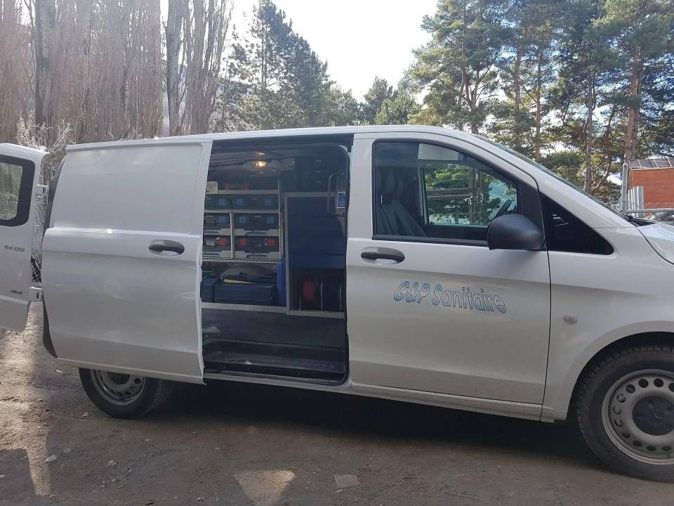 Fourgonnette de service blanche avec porte latérale ouverte, stationnée à l'extérieur ; intérieur visible avec étagères et équipement.