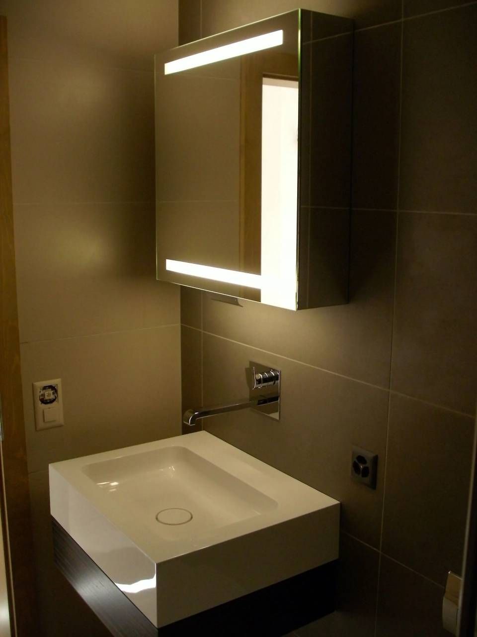 Salle de bains moderne avec lavabo blanc, armoire de toilette éclairée et murs carrelés gris.