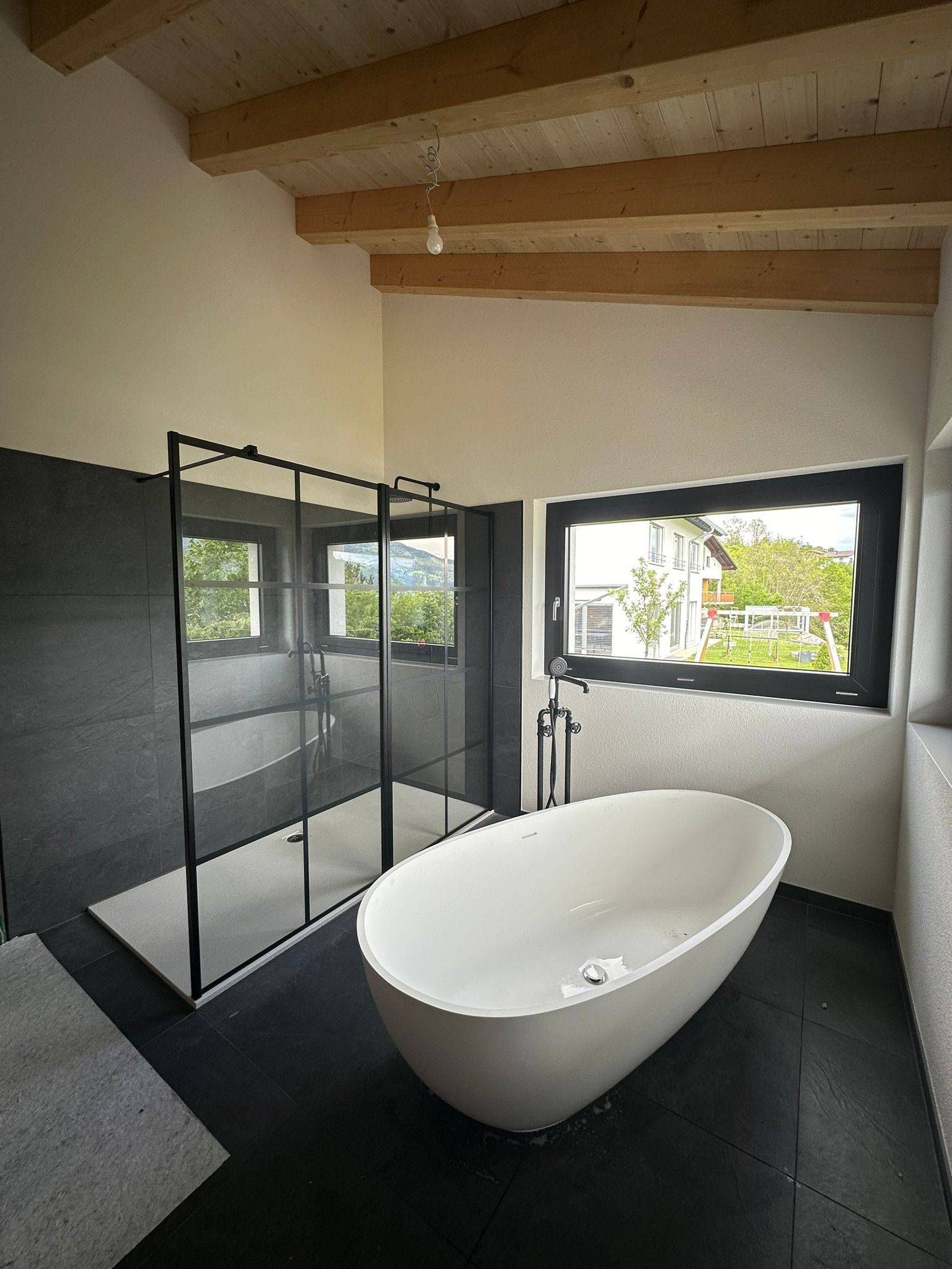 Salle de bains moderne avec baignoire îlot, douche en verre et poutres apparentes au plafond.