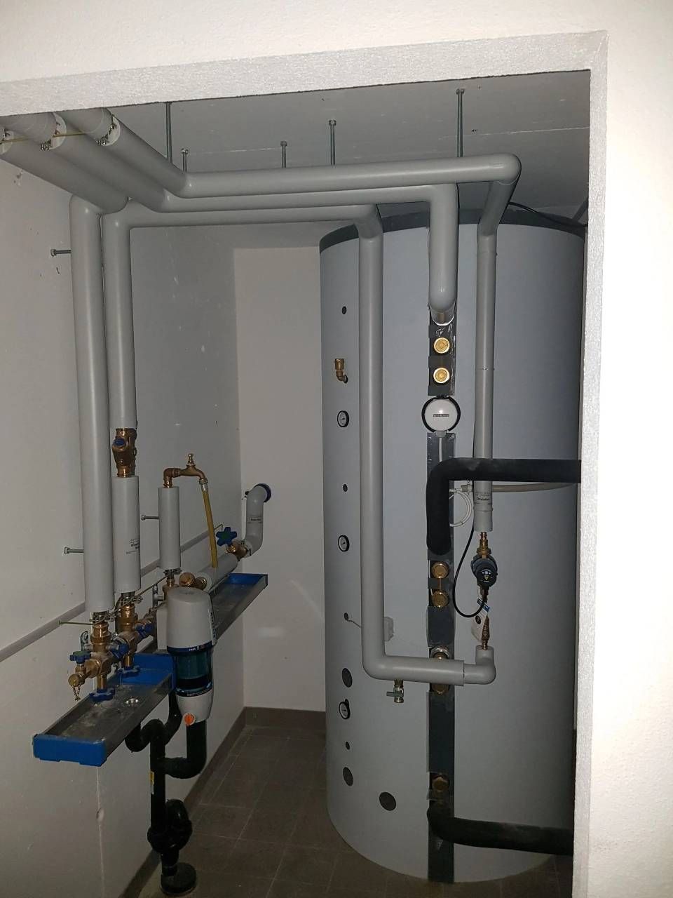 Local technique avec chauffe-eau vertical et tuyauterie raccordée ; murs blancs.