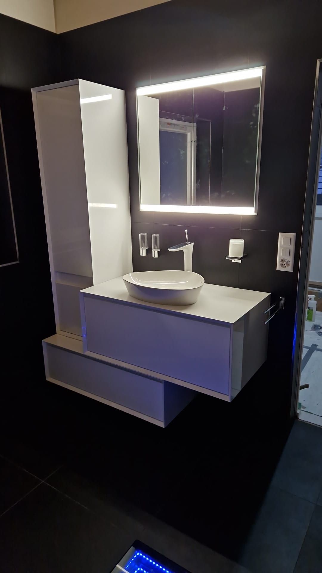 Un petit lavabo blanc en forme de seau avec un robinet noir, fixé sur un mur carrelé gris.