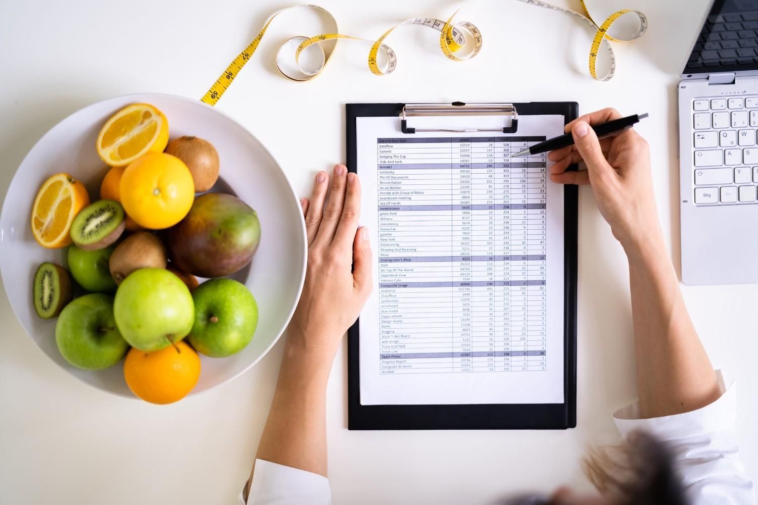 Manos rellenando un formulario en un portapapeles junto a un frutero y un ordenador portátil sobre un escritorio blanco.