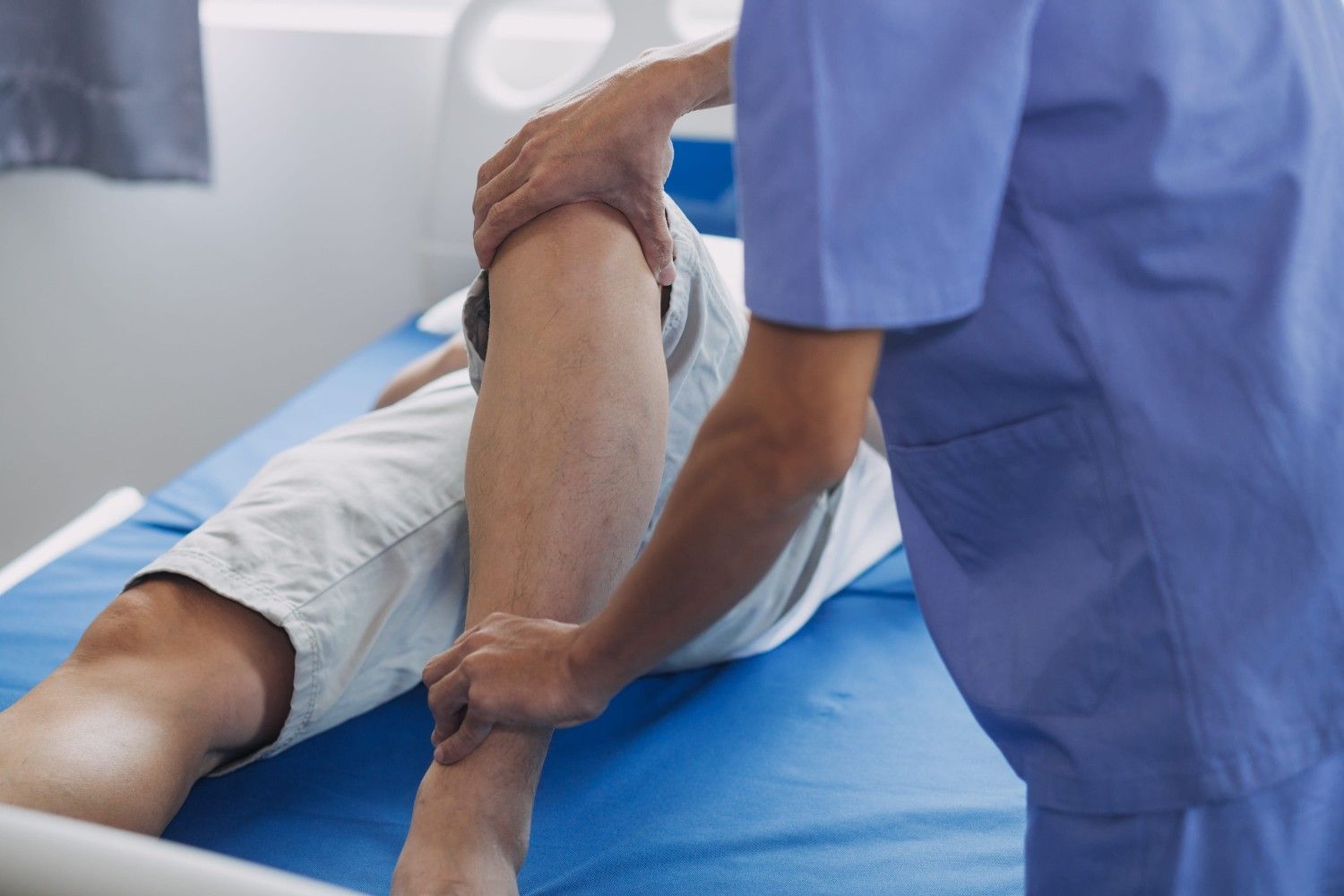 Un cuidador con uniforme azul sostiene la pierna de un paciente sobre una camilla de tratamiento en una sala de la clínica.