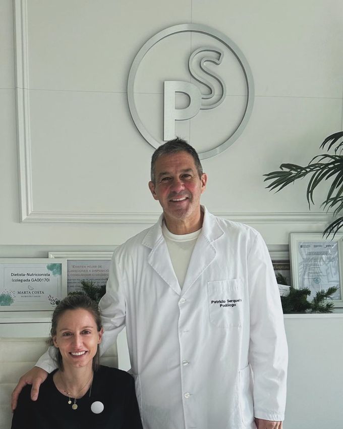 Dos personas sonriendo en la recepción de una clínica, una de ellas con bata blanca.
