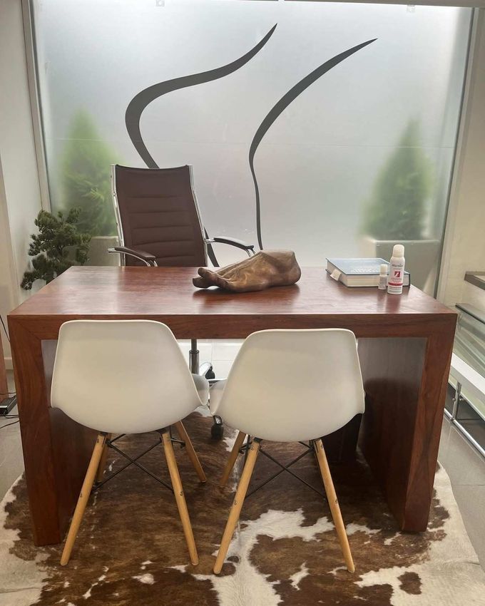 Pequeña oficina con un escritorio de madera, dos sillas blancas, una pared de vidrio esmerilado.