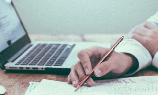 Persona escribiendo en un papel con un lápiz junto a un ordenador portátil.