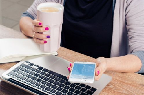 Mujer sentada en un escritorio con un ordenador portátil, sosteniendo un teléfono y una taza de café; uñas pintadas de rosa y morado.