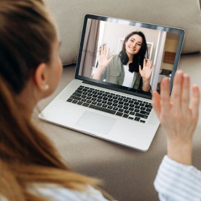 Mujer en la pantalla de un ordenador portátil saludando a otra persona que le devuelve el saludo; ambiente interior.