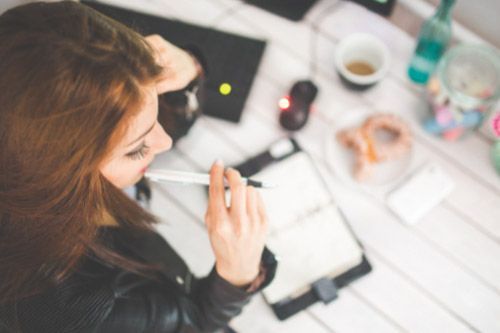 Mujer con bolígrafo en la boca, mirando un cuaderno abierto, trabajando en un escritorio con un ordenador portátil, café y rosquillas.