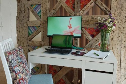 Un despacho en casa con un escritorio blanco, ordenador portátil, monitor y un jarrón con flores frente a una pared con vigas de madera.