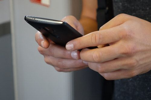 Manos de una persona sosteniendo un teléfono inteligente negro; un pulgar toca la pantalla, mientras que los demás dedos sostienen el dispositivo.