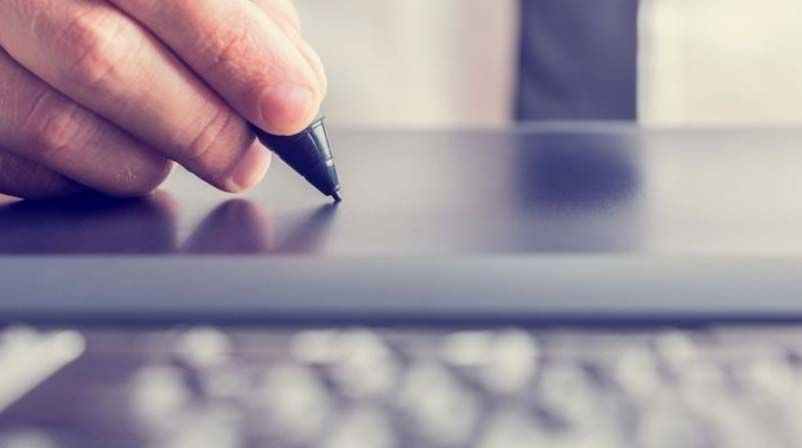 Una mano utiliza un lápiz óptico para escribir en una tableta digital negra; el teclado está en primer plano.