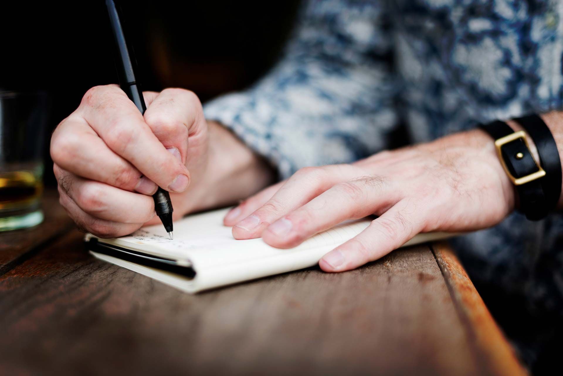 Persona escribiendo en una libreta sobre una mesa de madera; se ven el bolígrafo, la mano y el reloj de pulsera.