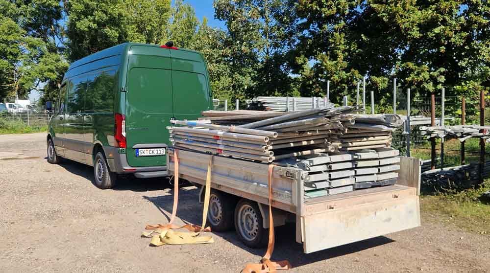 Ein grüner Transporter parkt neben einem Anhänger, der mit gestapelten Holzbrettern beladen ist, an einer Baustelle am Straßenrand