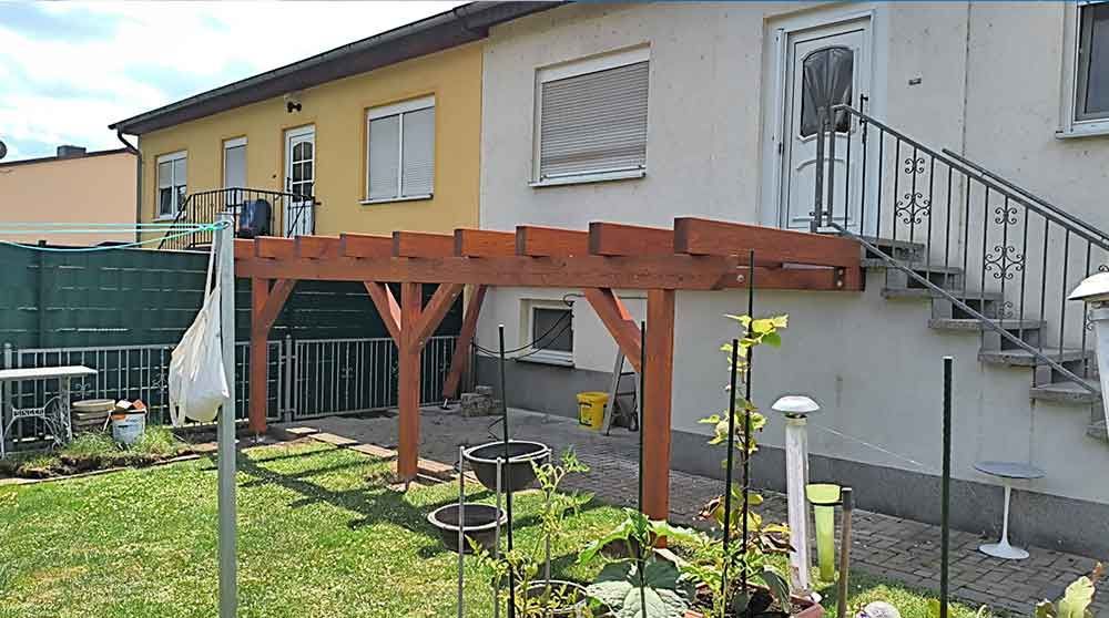 Hinterhof mit einer erhöhten Holzterrasse, die an ein gelbes Haus angebaut ist, und einer Außentreppe auf der rechten Seite