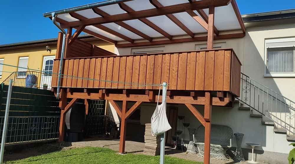 Holzterrasse mit Geländer und Überdachung am Haus, neben einer Rasenfläche und einer Treppe.