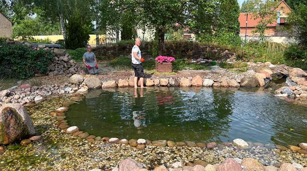 Zwei Personen stehen an einem kleinen Gartenteich, der von Felsen und Bäumen umgeben ist