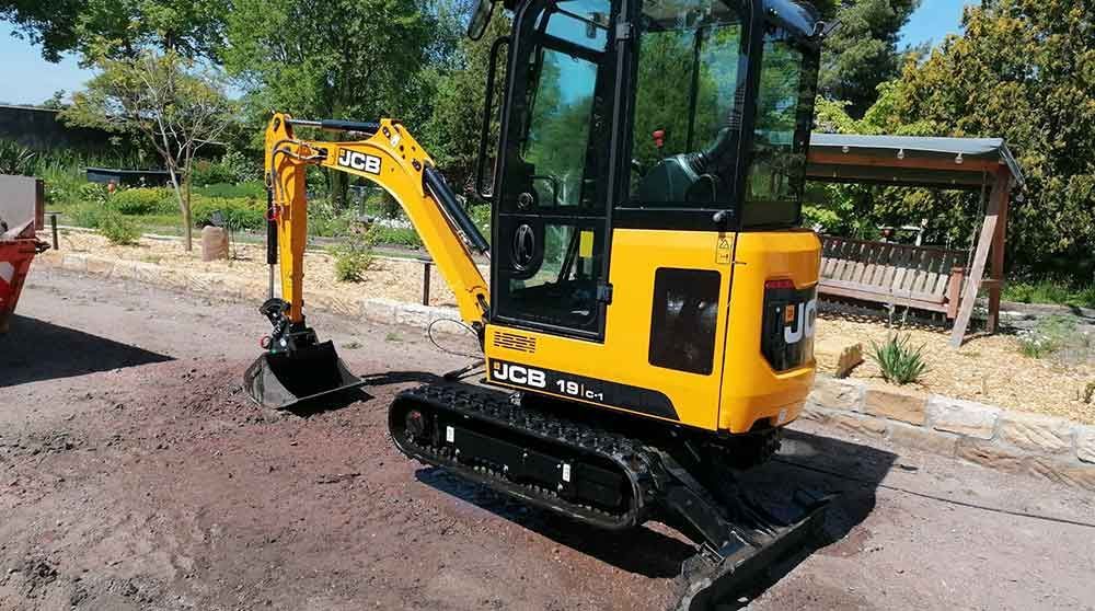 Gelber Minibagger auf Ketten, der draußen in der Nähe eines Gebäudes und von Bäumen Erde aushebt.