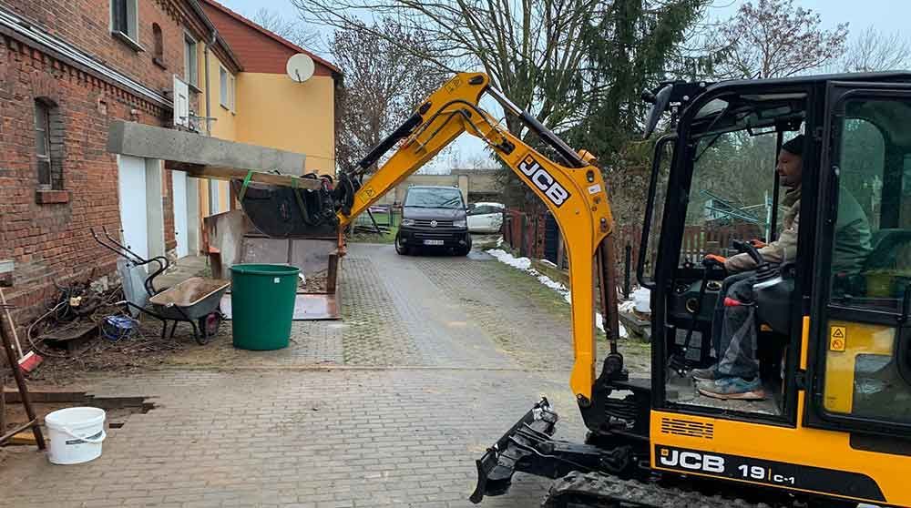 Gelber JCB-Bagger in einer Wohnstraße neben Backsteinhäusern und einer grünen Mülltonne