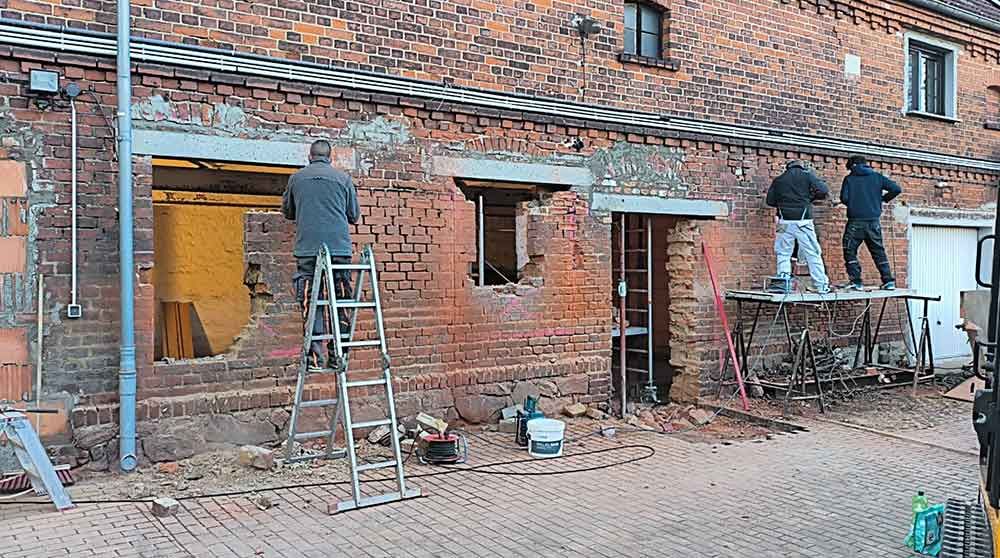 Arbeiter reparieren eine Ziegelmauer vor einem Backsteingebäude. Leitern, Werkzeuge und Bauschutt liegen auf dem Boden