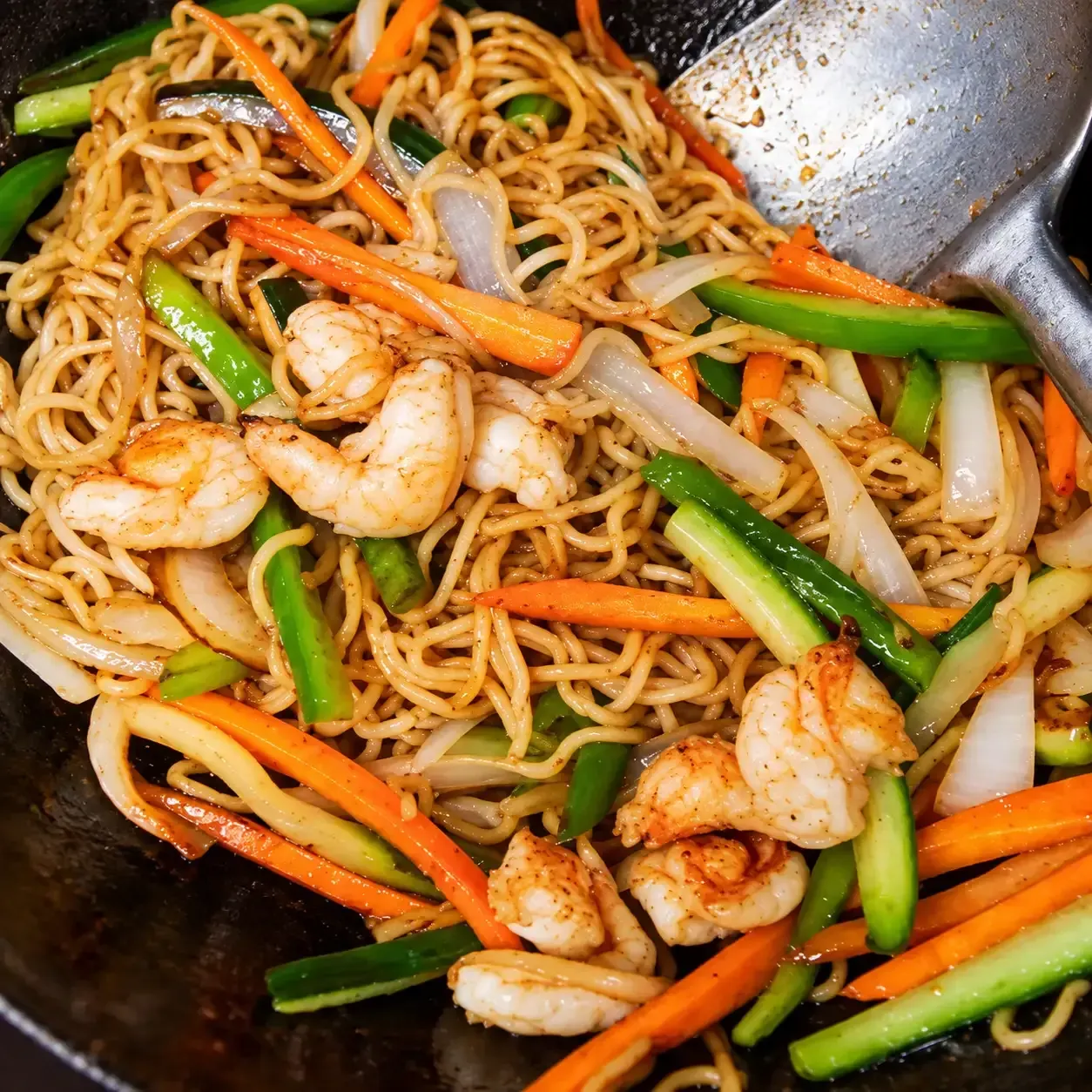 Fideos salteados con camarones y verduras en un wok, con pinzas a un lado.