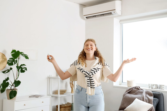 Une femme sourit, les bras tendus, sous un climatiseur dans une pièce lumineuse.