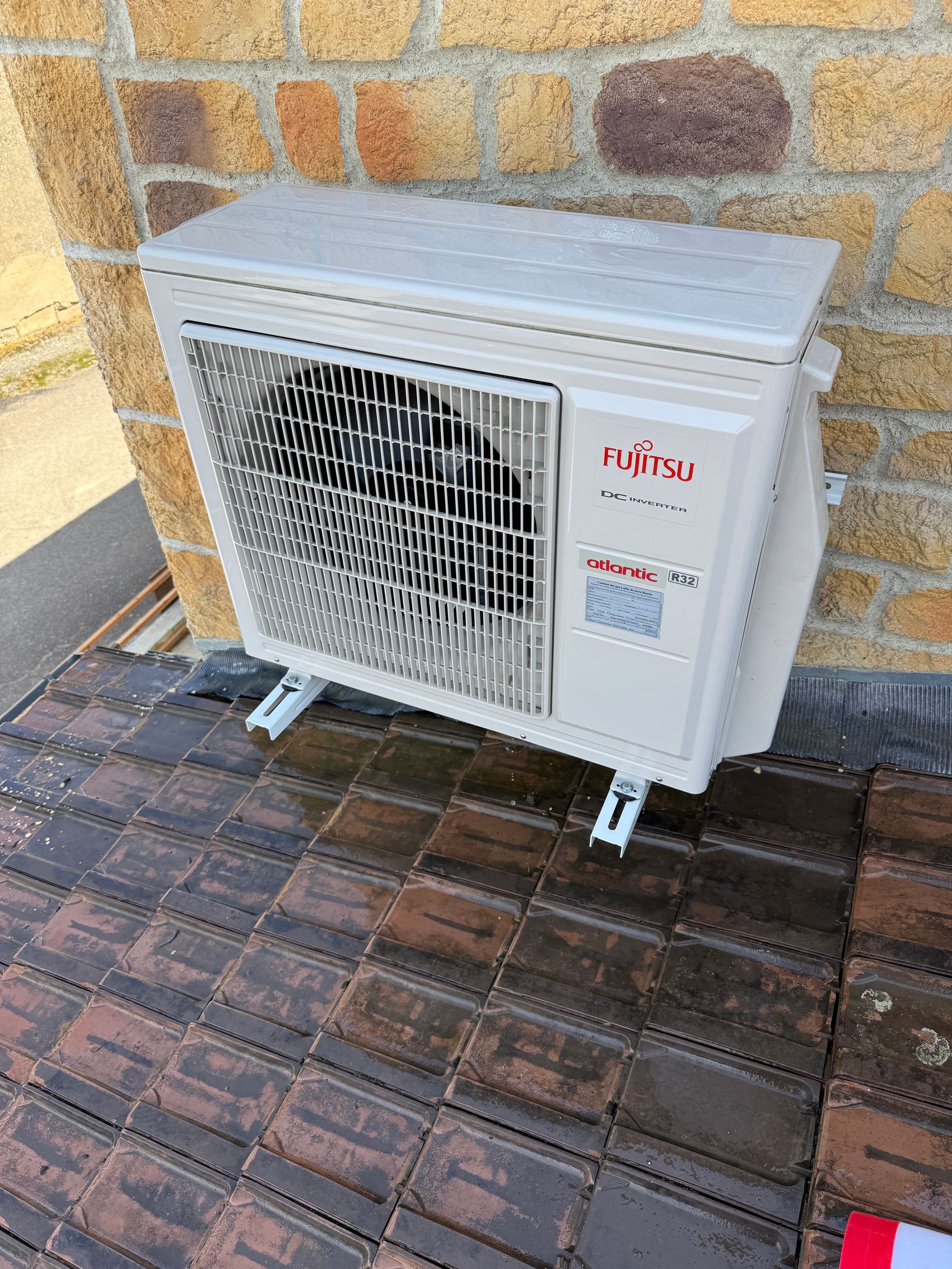 Unité de climatisation Fujitsu montée sur un toit en tuiles contre un mur de briques.