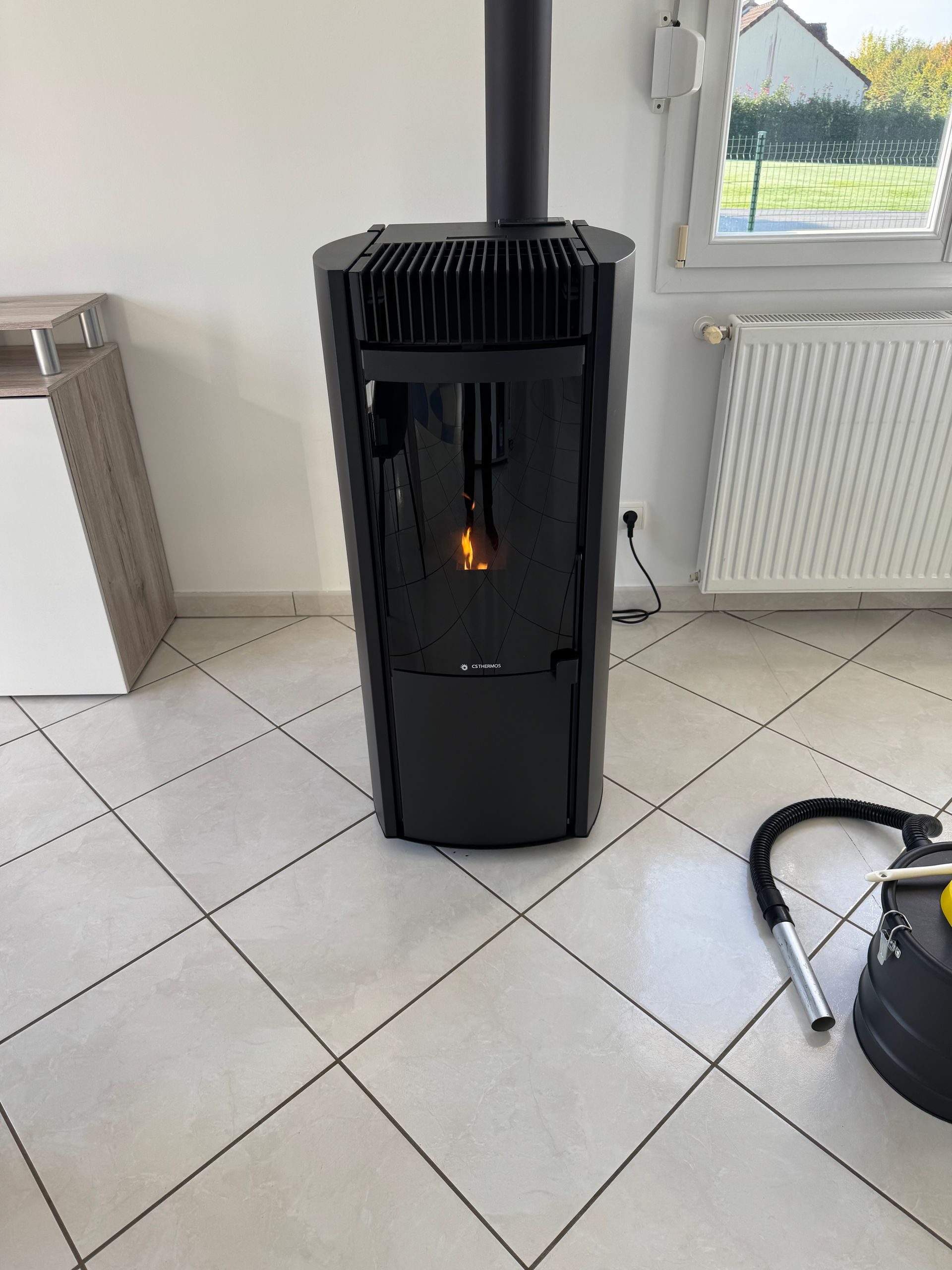 Poêle à granulés noir avec flamme, cheminée, sur sol carrelé, près de fenêtre et aspirateur.