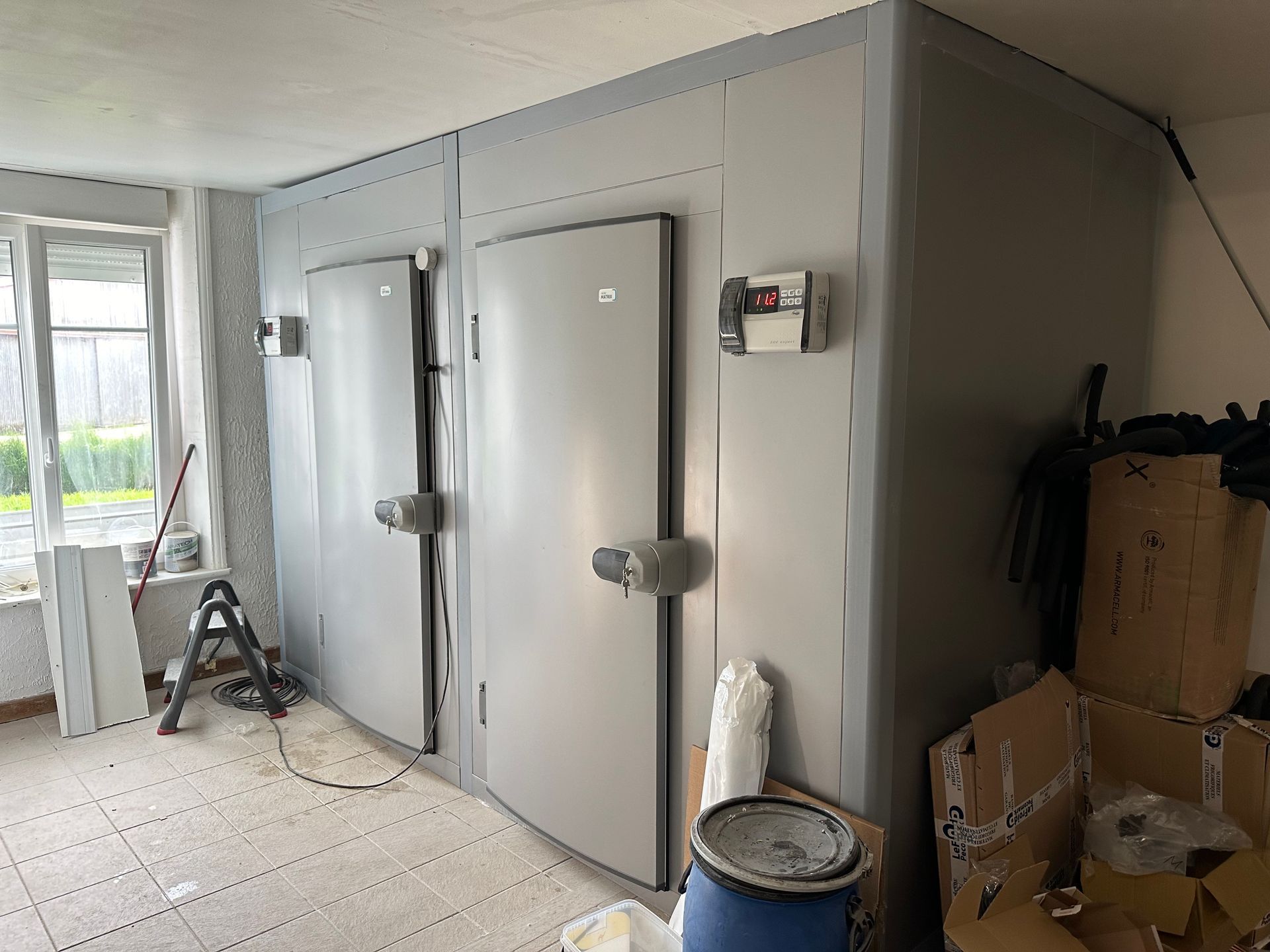 Deux chambres froides grises dans une pièce, avec portes, panneaux de commande et une fenêtre.