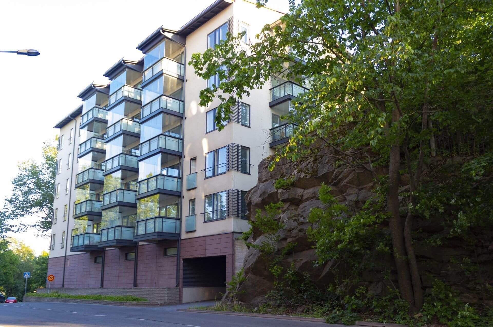 Kerrostalo lasiparvekkeineen kallioisen ja puiden peittämän kukkulan vieressä. Aurinkoinen päivä.