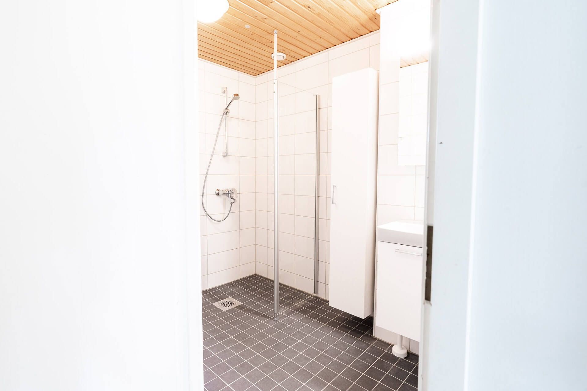 Kylpyhuoneen sisustus, jossa suihku, valkoiset kaapit, tumma laattalattia ja puinen katto.