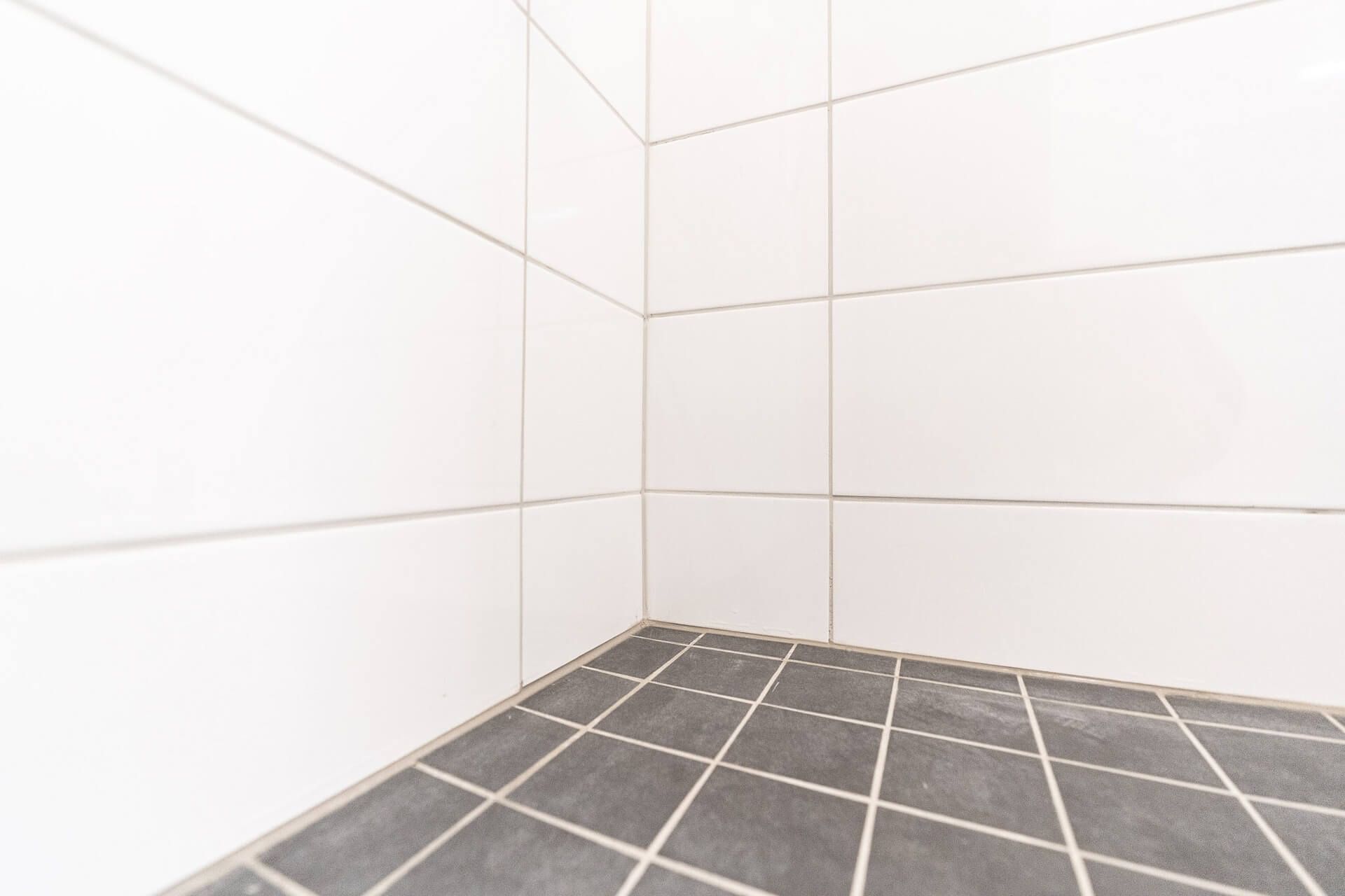 Laatoitetun kylpyhuoneen nurkka, jossa on valkoiset seinälaatat ja harmaat lattialaatat.