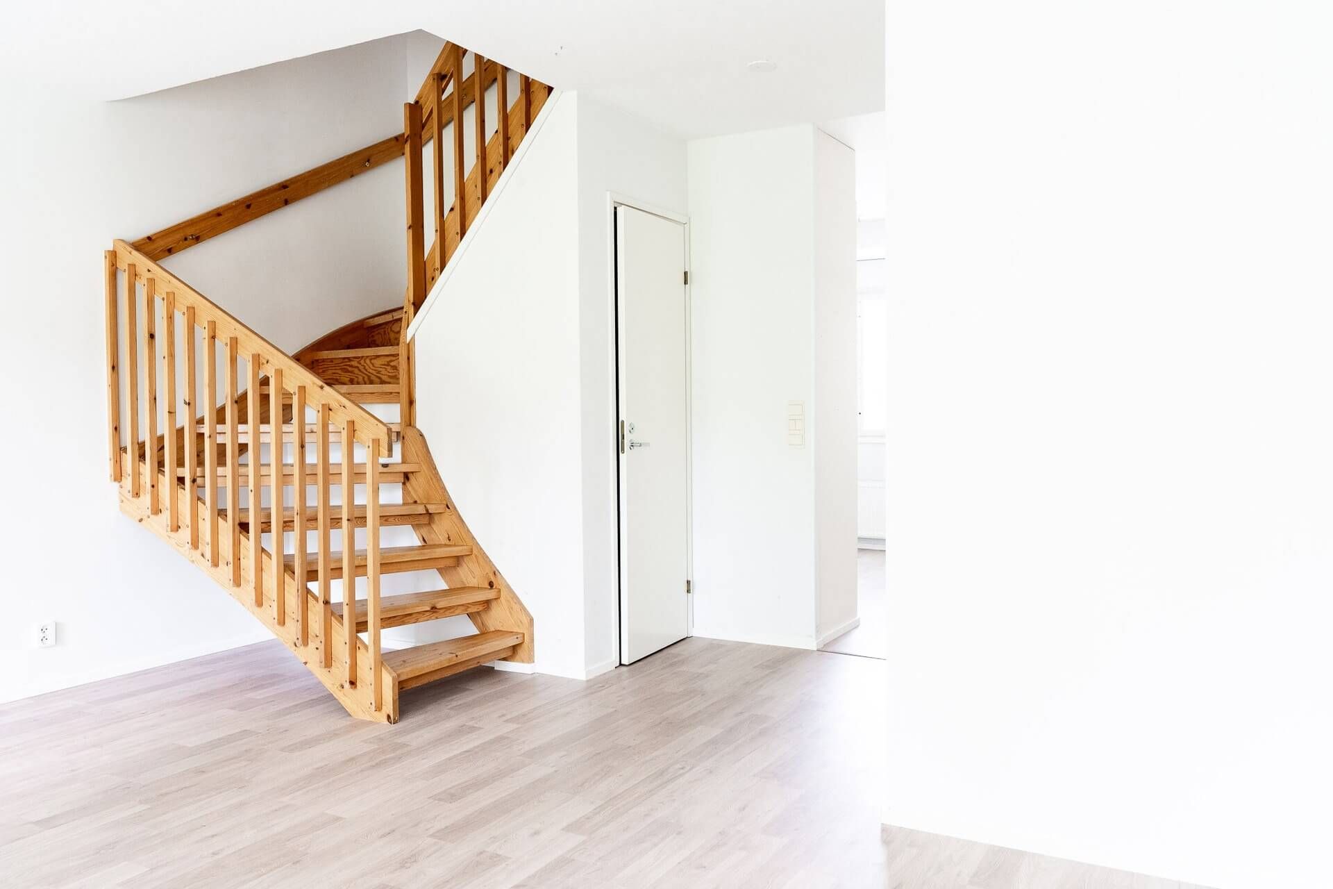 Puinen portaikko valkoisessa huoneessa, jonka ylhäällä näkyy ovi.