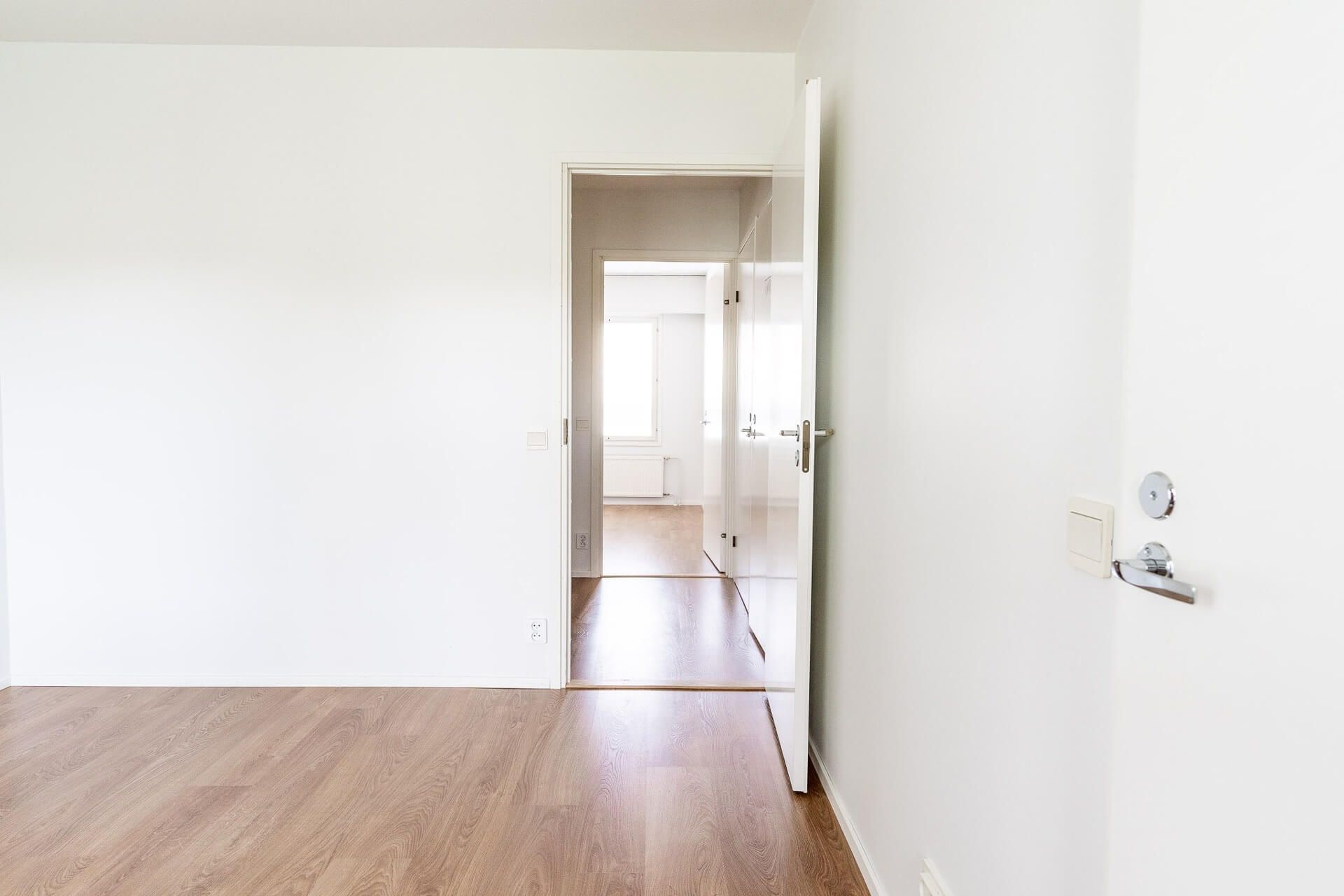 Avoin oviaukko paljastaa käytävän, joka johtaa ikkunalle. Huoneessa on vaalea puulattia ja valkoiset seinät.