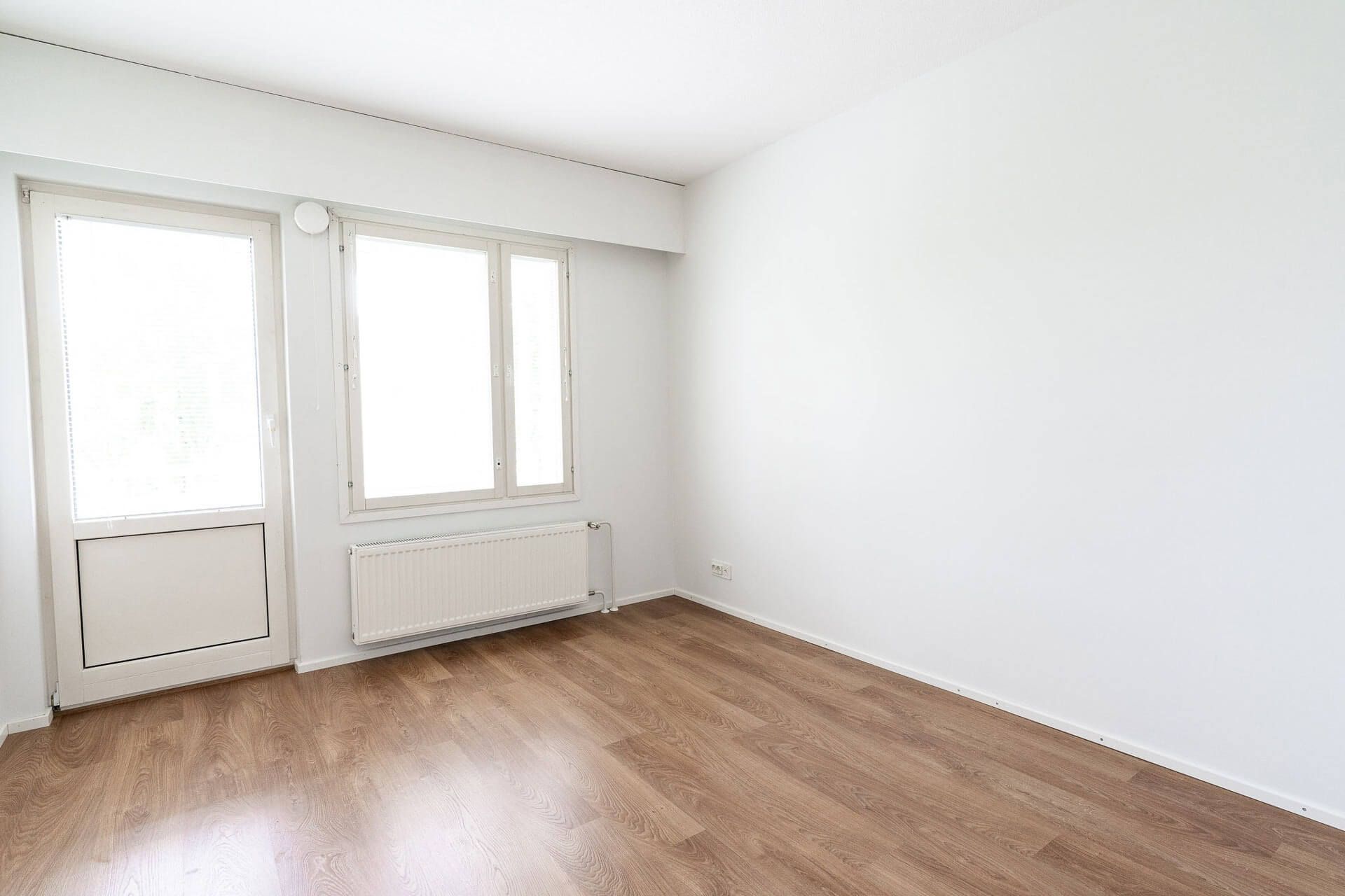 Tyhjä huone, jossa on puujäljitelmälattia ja valkoiset seinät. Ikkuna ja ovi ovat vasemmalla puolella.